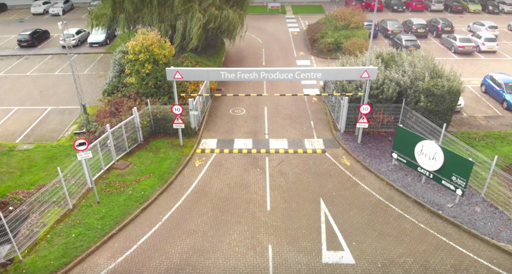 Widok z góry na wjazd do The Fresh Produce Centre z szlabanem i znakami drogowymi, parking w tle, w pochmurny dzień. Widoczny znak 'Fresh' Gate 3.