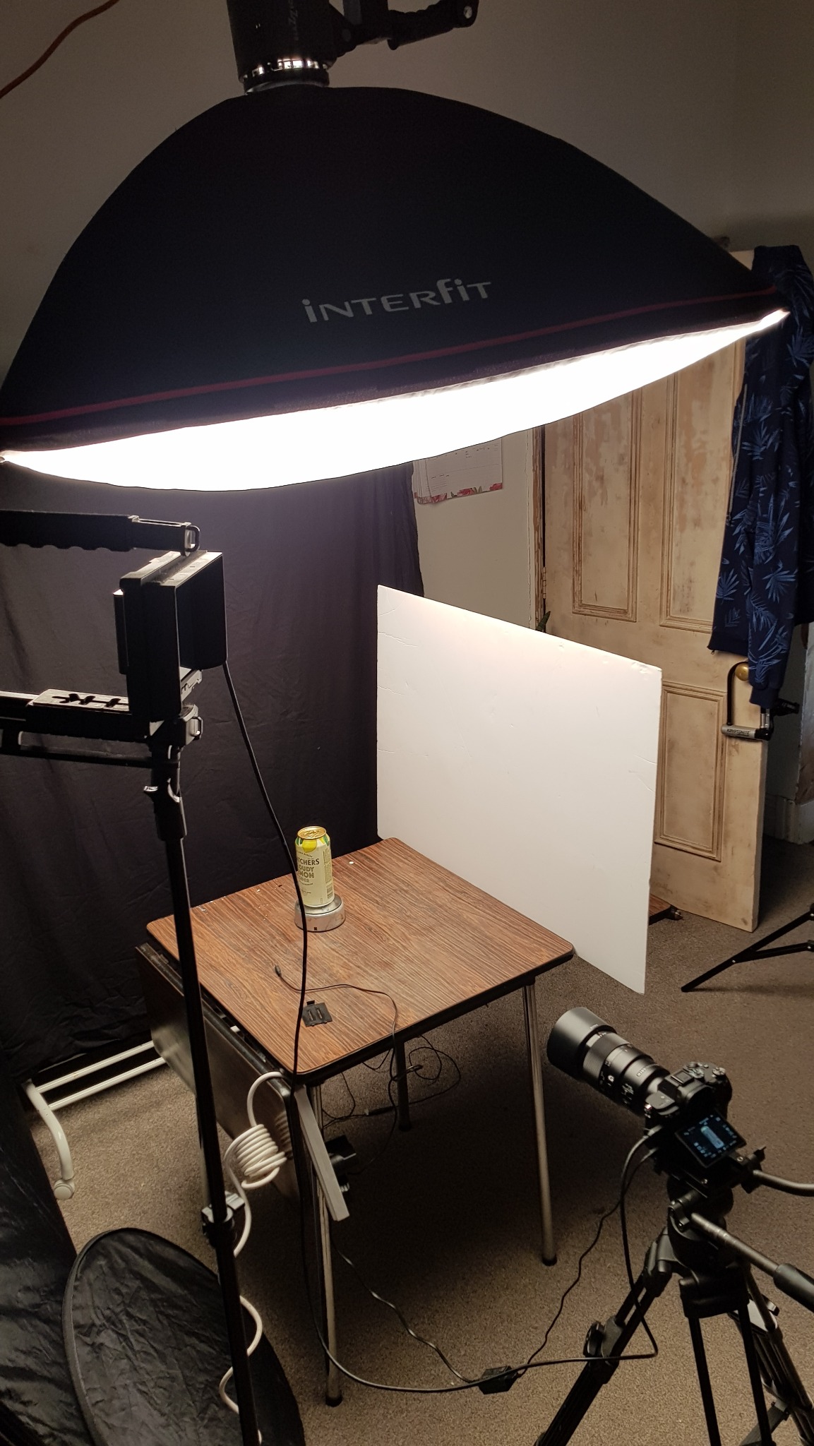 Studio fotograficzne z ustawionym oświetleniem Interfit, aparatem na statywie i produktem (puszka) na stole, gotowe do sesji produktowej. Białe tło i czarne zasłony.