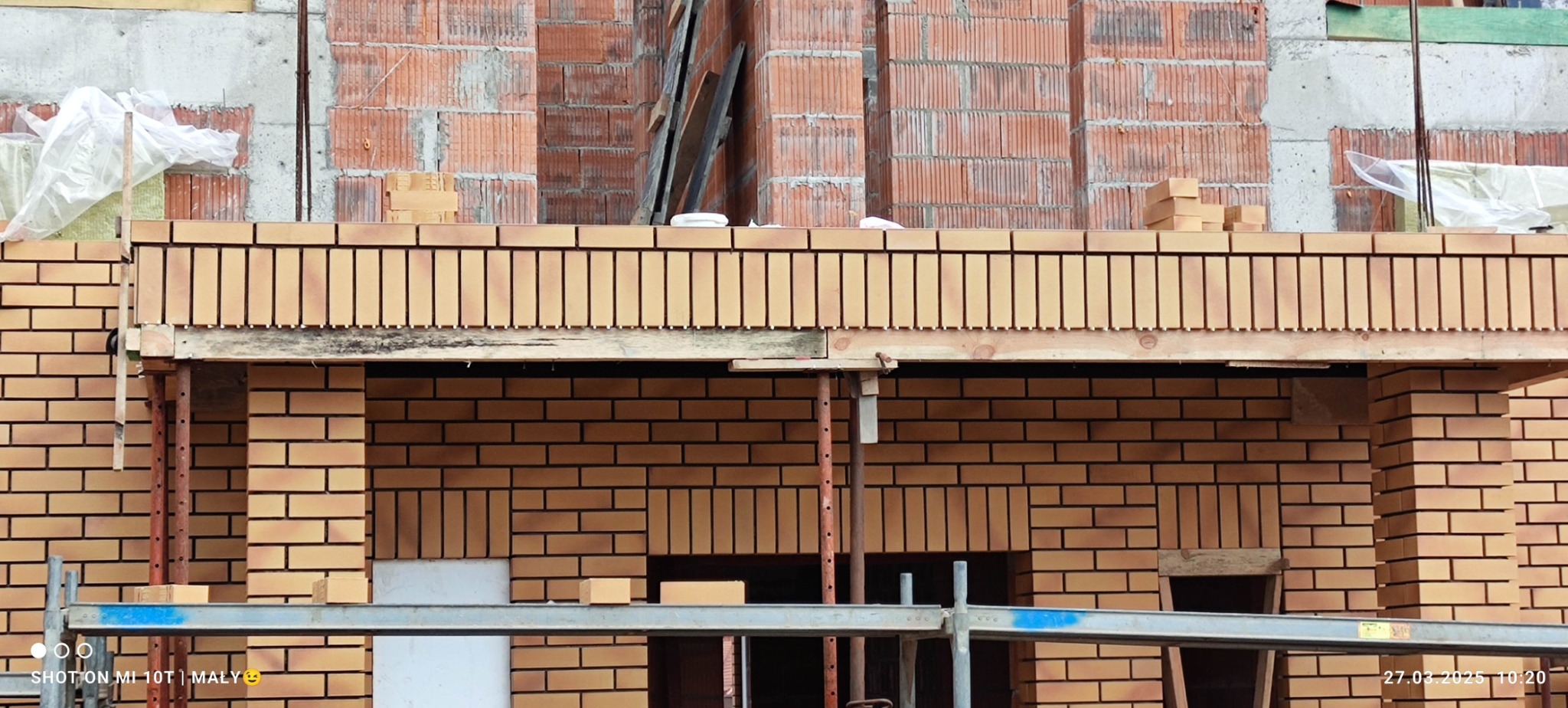 Murowana elewacja budynku w trakcie budowy, widoczne rusztowania i cegły klinkierowe. Częściowo ocieplony fragment ściany.