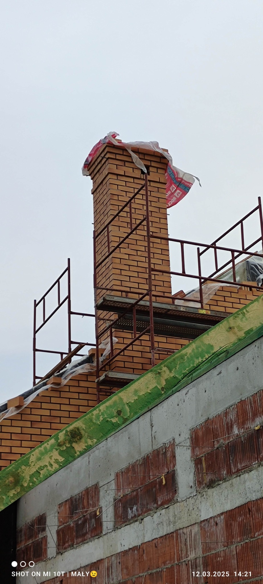 Wznoszenie komina z cegły klinkierowej na dachu budynku w trakcie budowy, widoczne rusztowanie i zabezpieczenie komina folią. Prace murarskie w toku.