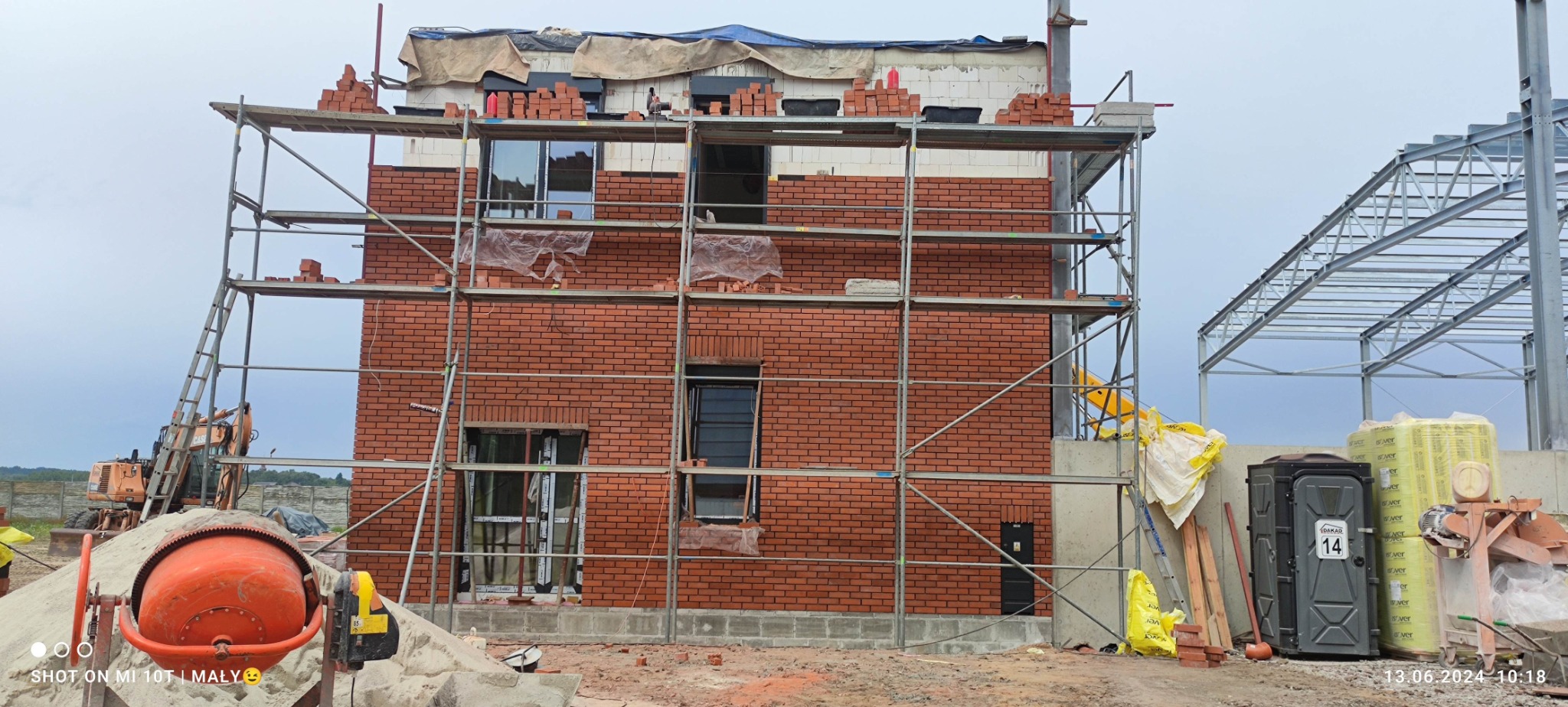 Budowa domu z cegły: wznoszenie dwukondygnacyjnej elewacji z czerwonej cegły, widoczne rusztowania i materiały budowlane wokół placu budowy.