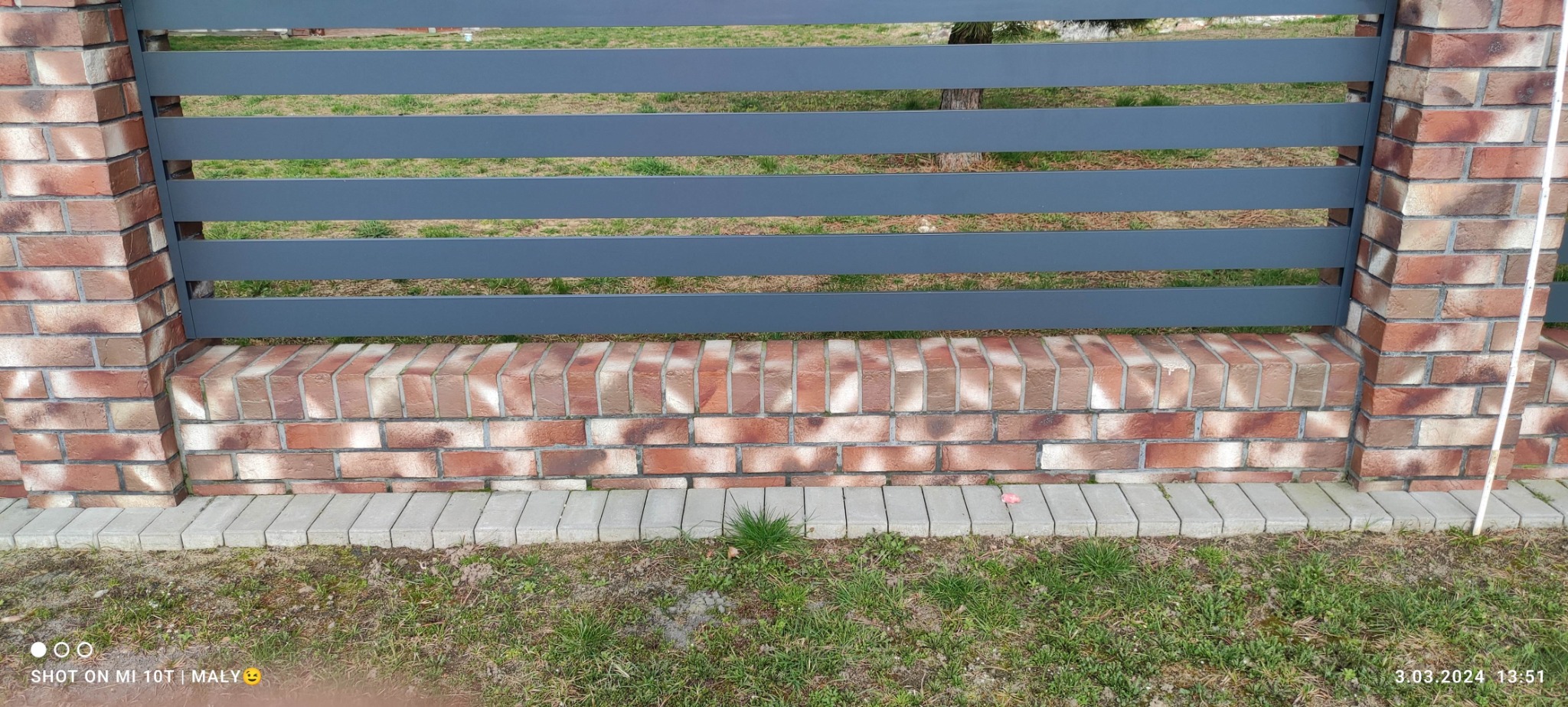 Nowoczesne ogrodzenie z cegły klinkierowej i szarych, poziomych przęseł. Widoczne detale murarskie i trawa przy podstawie. Kąt ujęcia z dołu podkreśla solidność konstrukcji.