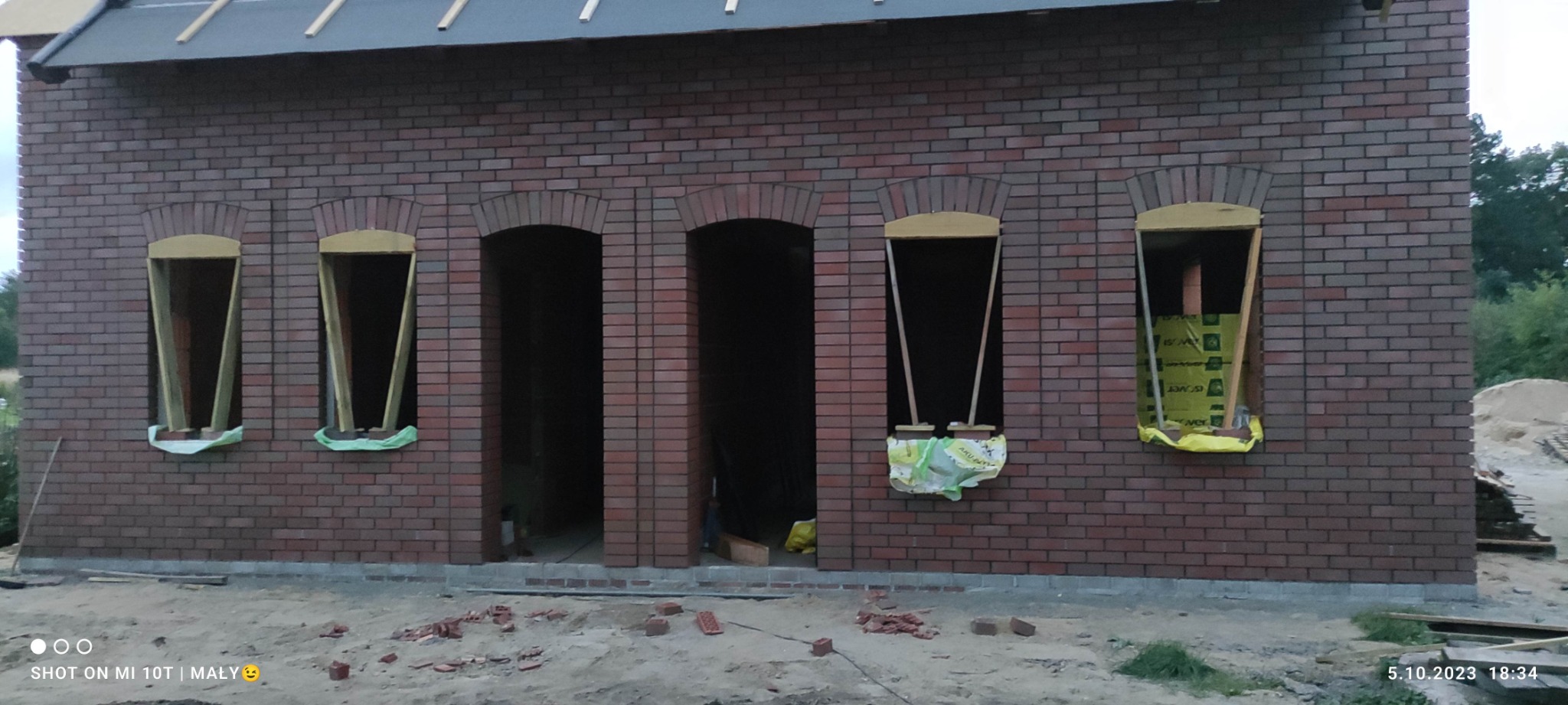 Budynek z cegły klinkierowej w trakcie budowy, widoczne otwory okienne z drewnianą konstrukcją, częściowo zabezpieczone folią. Surowy stan, widoczne elementy budowlane.