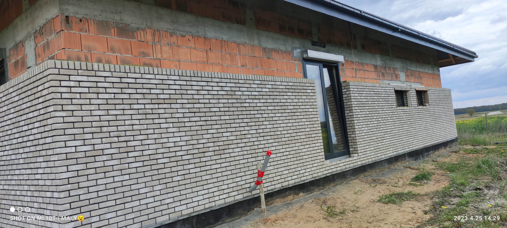 Fragment elewacji budynku w trakcie prac, z widocznym murem z cegły klinkierowej i oknem z roletą. W tle pole i niebo.