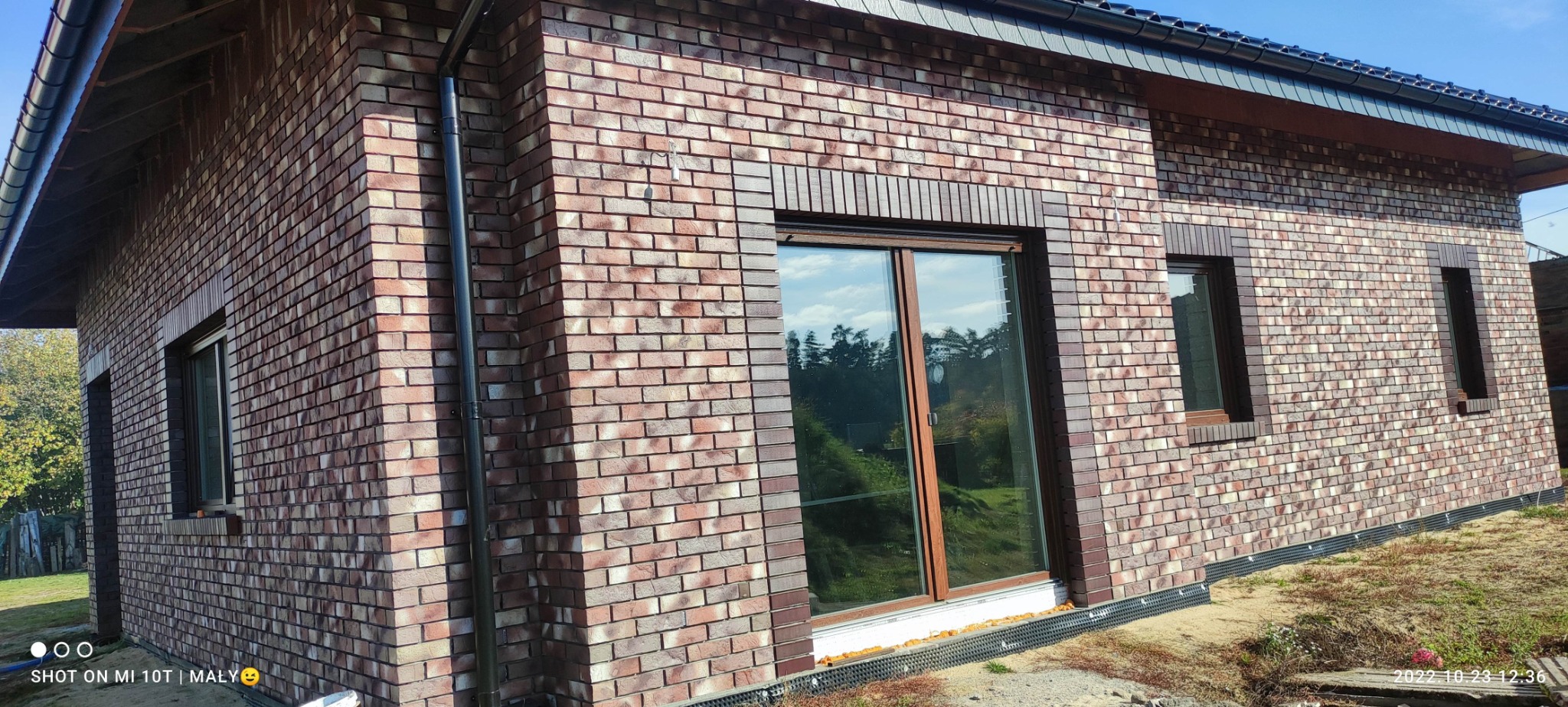 Elewacja budynku wykończona cegłą klinkierową w odcieniach czerwieni i brązu. Widoczne okna z drewnianą stolarką i czarny system orynnowania. Detal wykończenia.