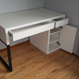 Paweł Zapasek - Nowoczesne biurko na metalowych nogach z białymi frontami, otwartymi szufladami i szafką z półkami, na tle drewnianej podłogi i grzejnika.