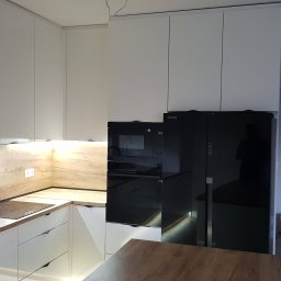 Paweł Zapasek - Nowoczesna kuchnia z białymi szafkami i drewnianym blatem, czarny sprzęt AGD, oświetlenie LED pod szafkami. Widoczny fragment blatu roboczego i okno z żaluzjami.