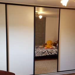 Paweł Zapasek - Biała szafa wnękowa z przesuwnymi drzwiami i czarnymi obramowaniami, w środkowym lustrze odbija się pokój z łóżkiem i pluszową zabawką.