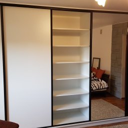 Paweł Zapasek - Biała szafa wnękowa z czarnymi obramowaniami i lustrzanymi drzwiami, odbijająca pokój z łóżkiem. Jedna sekcja szafy otwarta, ukazująca puste półki.