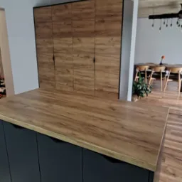 Nowoczesna szafa wnękowa z drewnianymi frontami i czarnymi uchwytami, widoczna z perspektywy blatu kuchennego, z elementami jadalni w tle.