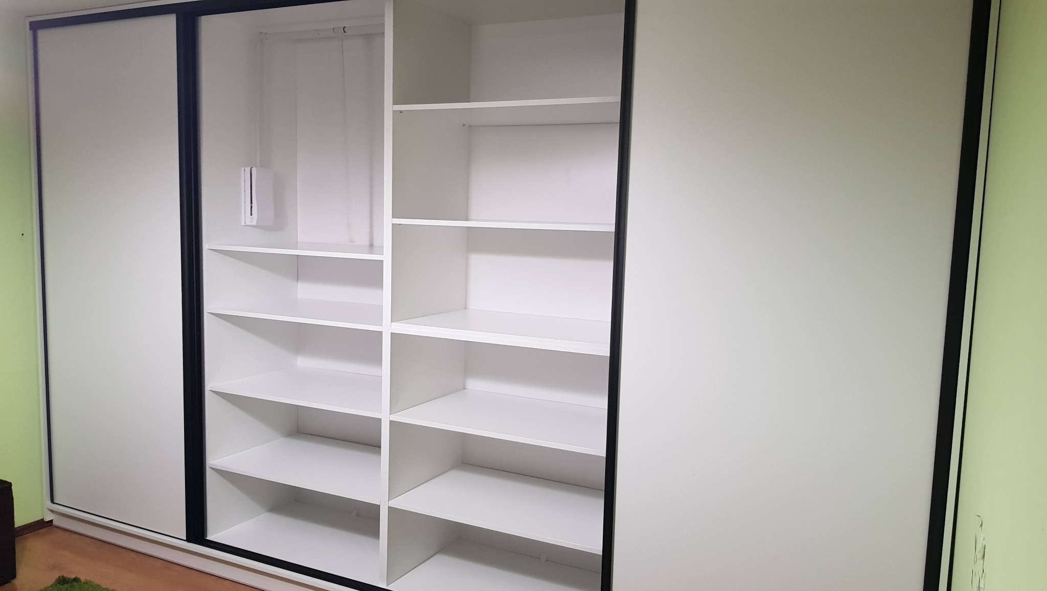 Biała szafa wnękowa z przesuwnymi drzwiami i czarnymi obramowaniami, z widocznymi półkami w otwartej części. Minimalistyczny design.