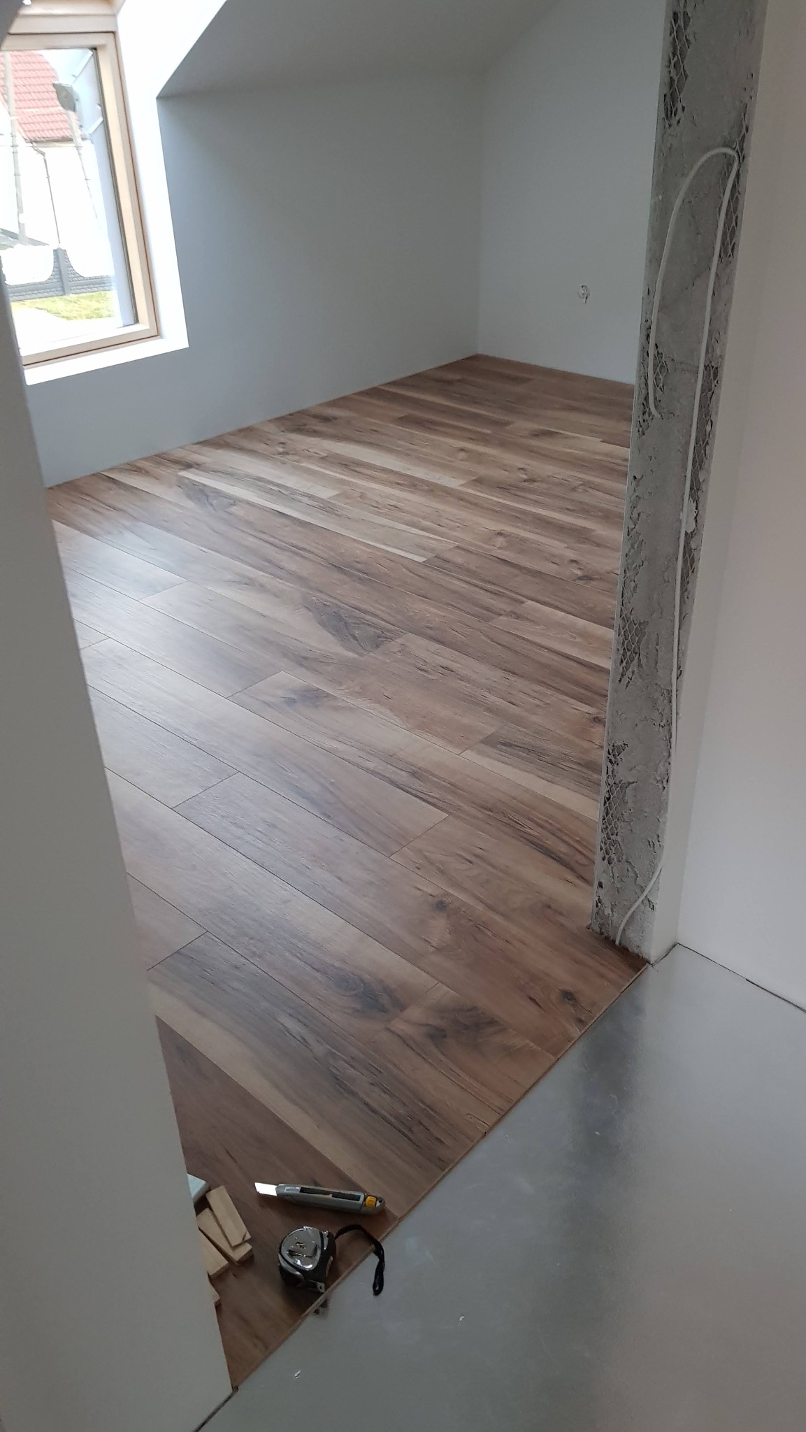 Ukończone układanie paneli podłogowych w pokoju na poddaszu, widoczne narzędzia pracy: miarka i nóż. Część podłogi wyłożona folią.