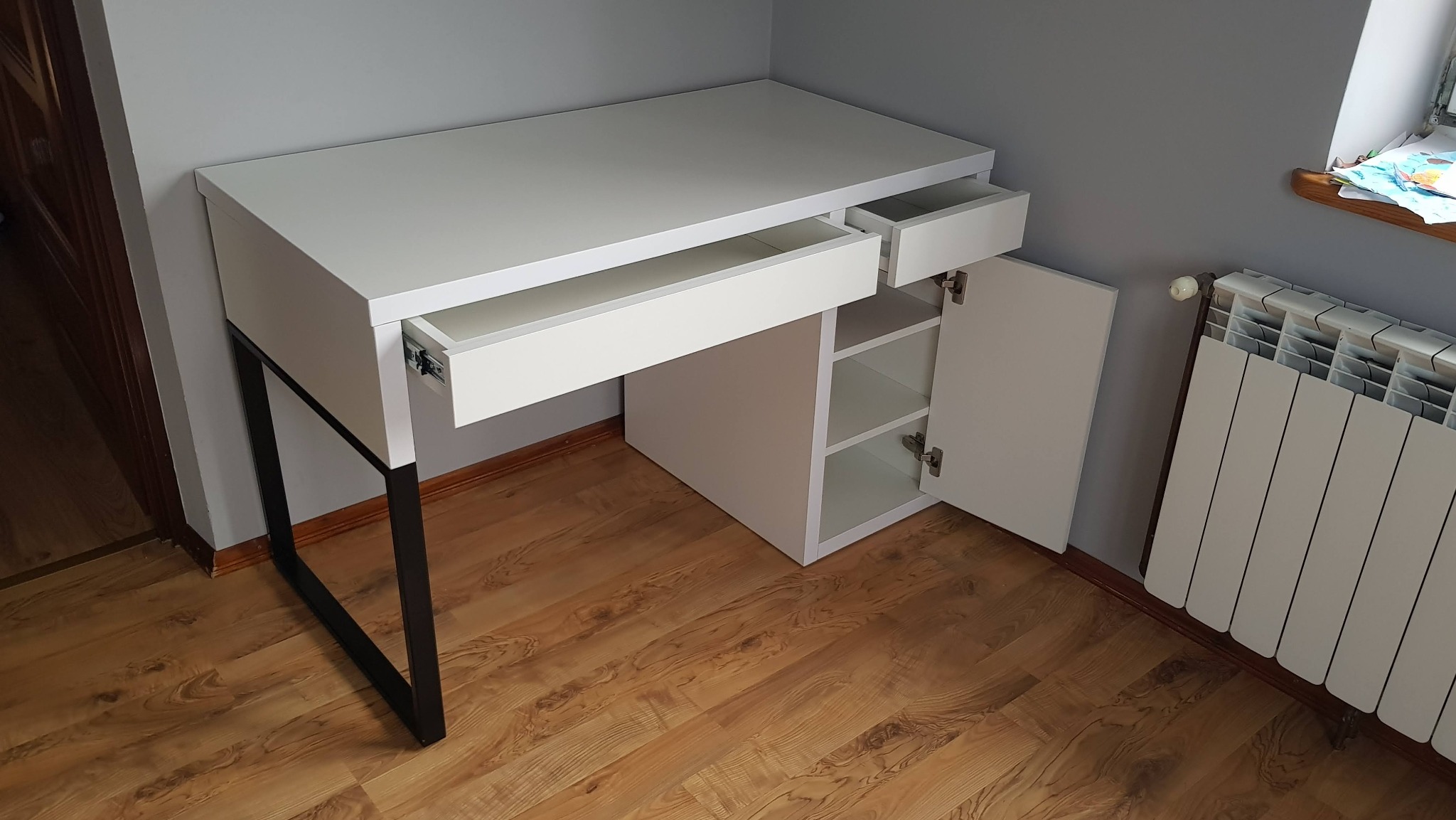 Nowoczesne biurko na metalowych nogach z białymi frontami, otwartymi szufladami i szafką z półkami, na tle drewnianej podłogi i grzejnika.