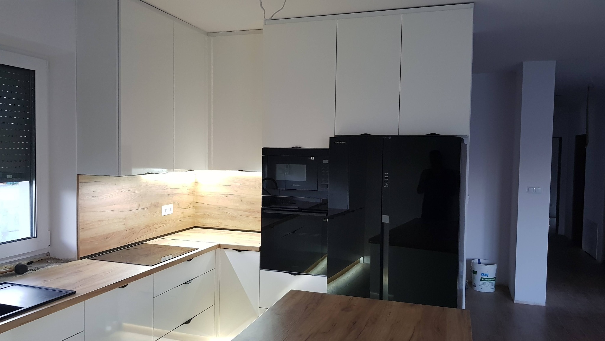 Nowoczesna kuchnia z białymi szafkami i drewnianym blatem, czarny sprzęt AGD, oświetlenie LED pod szafkami. Widoczny fragment blatu roboczego i okno z żaluzjami.