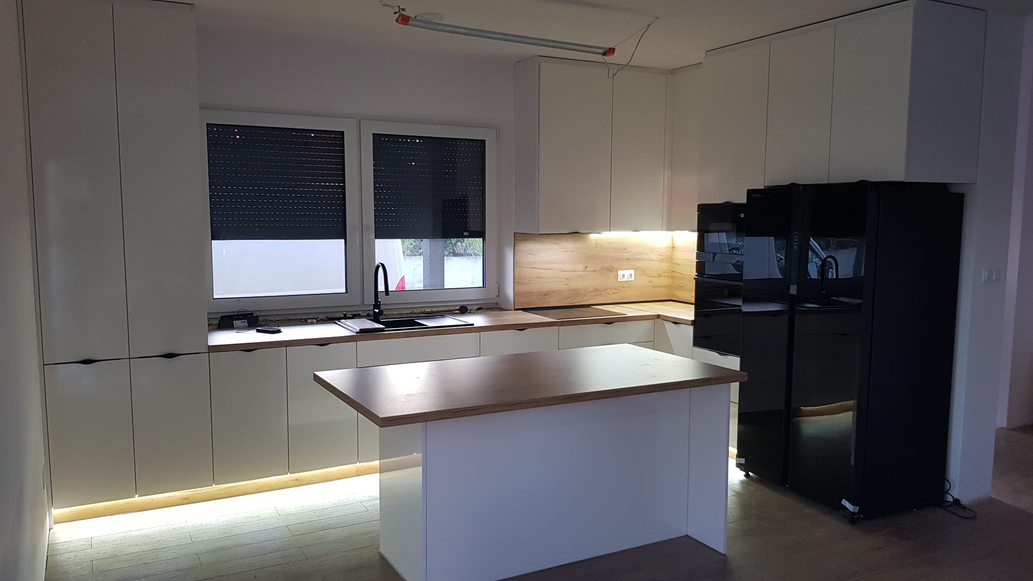 Jasna kuchnia z wyspą i czarną lodówką. Blat drewniany, szafki białe z podświetleniem. Okno z roletami, oświetlenie podszafkowe.