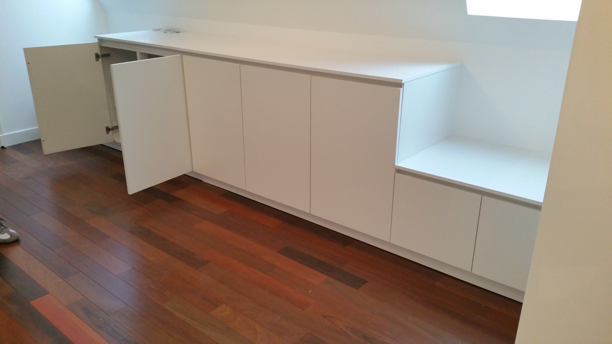 Biała zabudowa wnękowa ze skrzydłowymi drzwiami, drewniana podłoga. Otwarta szafka z półką po prawej stronie. Jedne drzwi szafki są otwarte.