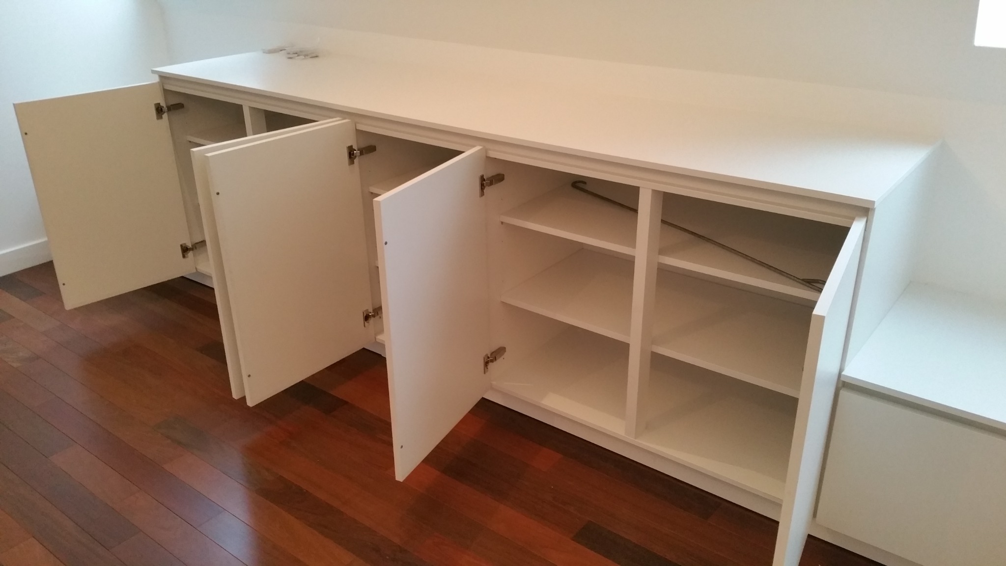 Biała szafa wnękowa z otwartymi drzwiczkami, widoczne półki i zawiasy, na tle parkietu. Minimalistyczny design i funkcjonalne rozwiązanie do przechowywania.