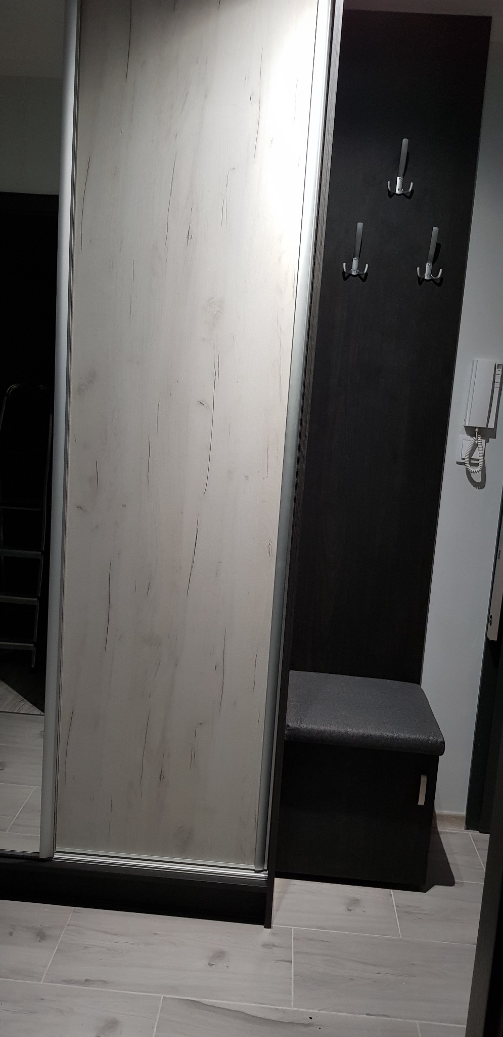 Nowoczesna szafa wnękowa z przesuwnymi drzwiami w kolorze jasnego drewna, połączona z czarnym panelem z wieszakami i siedziskiem. Minimalistyczny design.