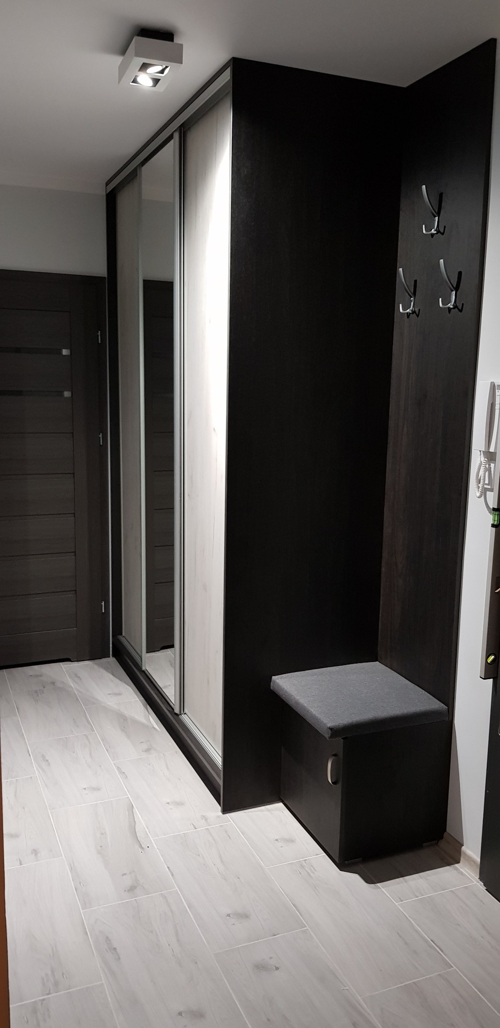 Nowoczesna szafa wnękowa z lustrzanymi drzwiami przesuwnymi i wbudowanym siedziskiem z wieszakami w przedpokoju. Widoczne panele podłogowe i drzwi.