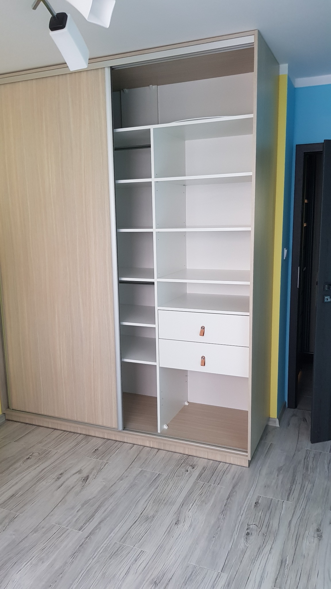 Otwarta szafa wnękowa z przesuwnymi drzwiami, z półkami i szufladami w Sokołowie Małopolskim. Jasne drewno na drzwiach, białe wnętrze i panele na podłodze.