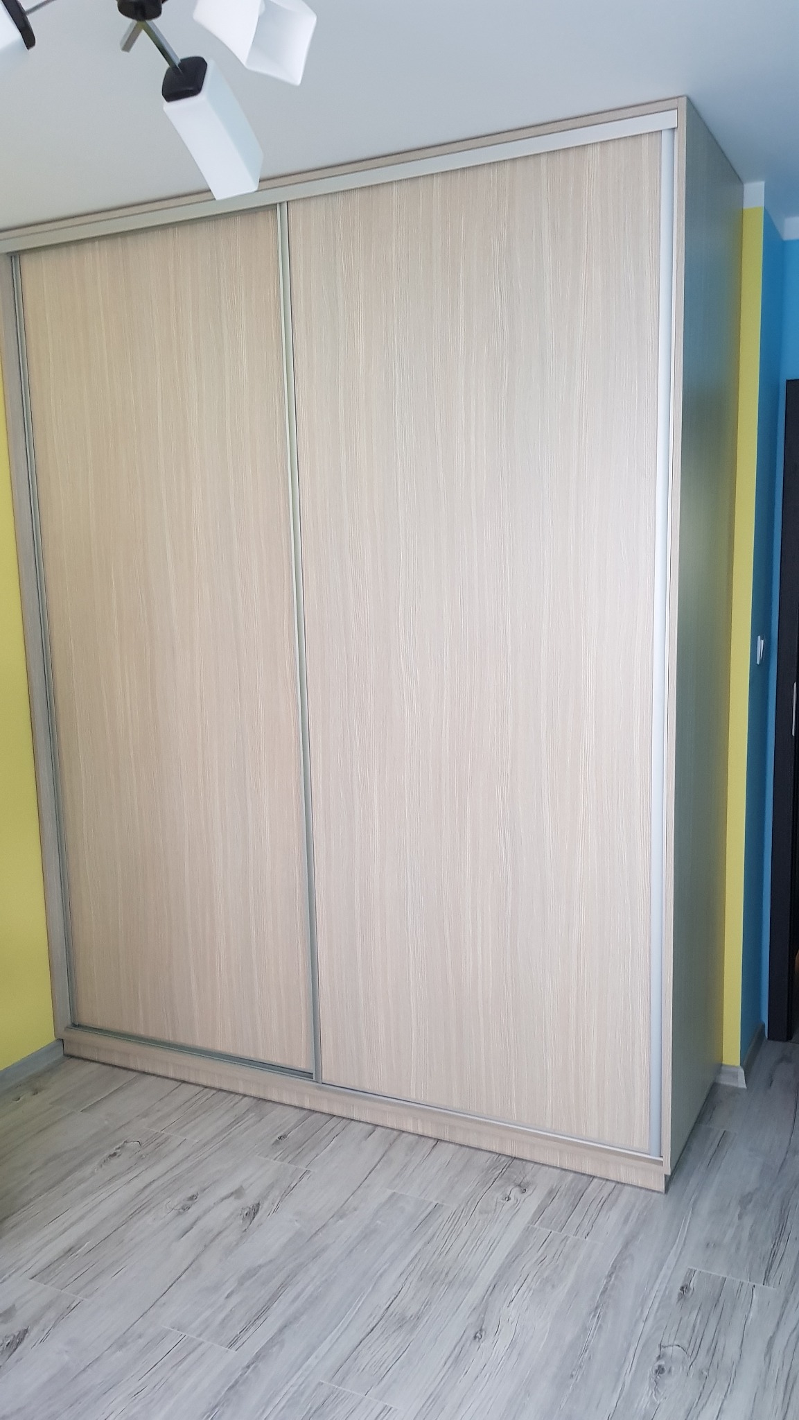 Jasna szafa wnękowa z przesuwnymi drzwiami, widoczna na tle parkietu i kolorowych ścian, oświetlona światłem z lampy sufitowej.