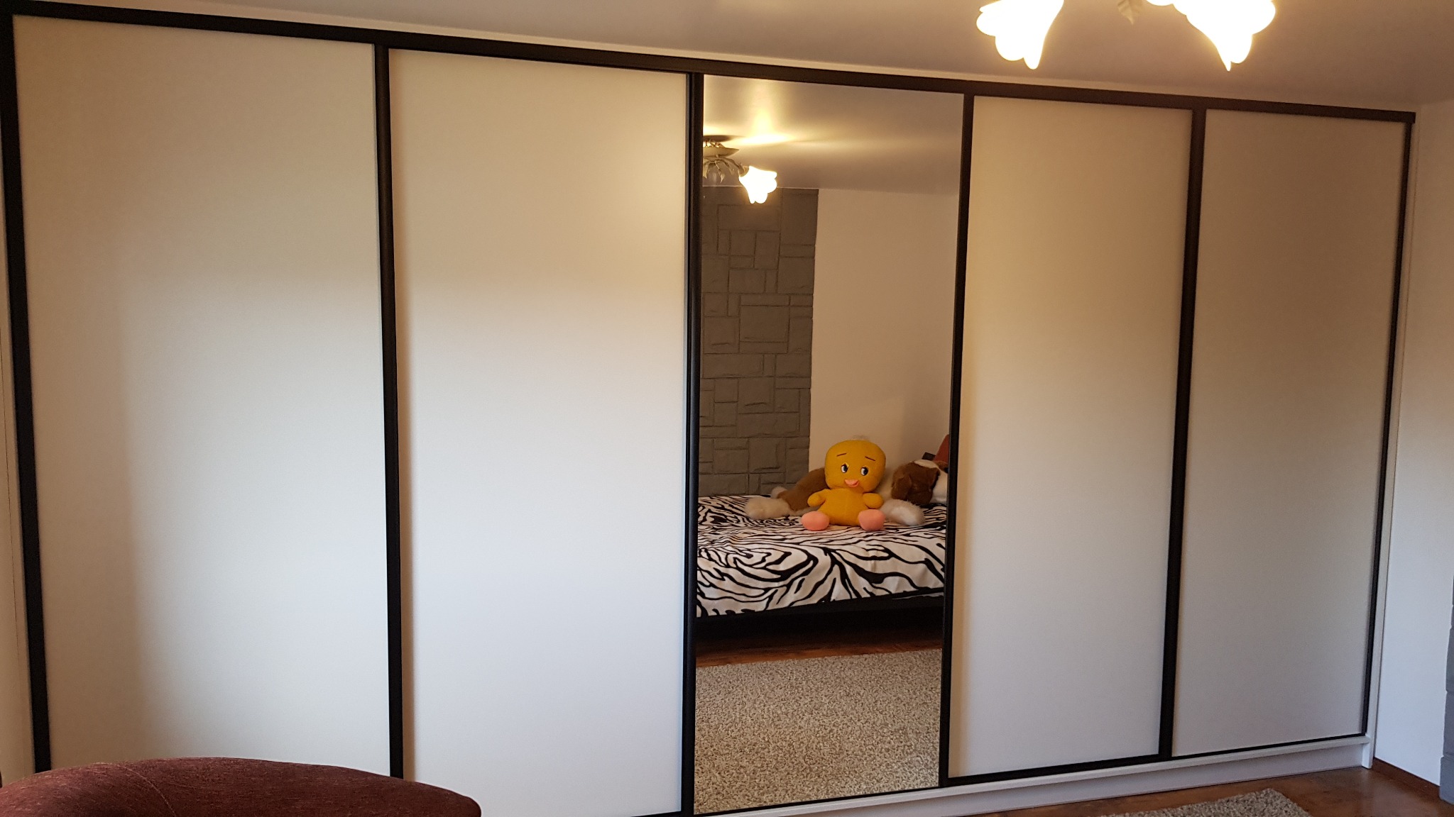 Biała szafa wnękowa z przesuwnymi drzwiami i czarnymi obramowaniami, w środkowym lustrze odbija się pokój z łóżkiem i pluszową zabawką.