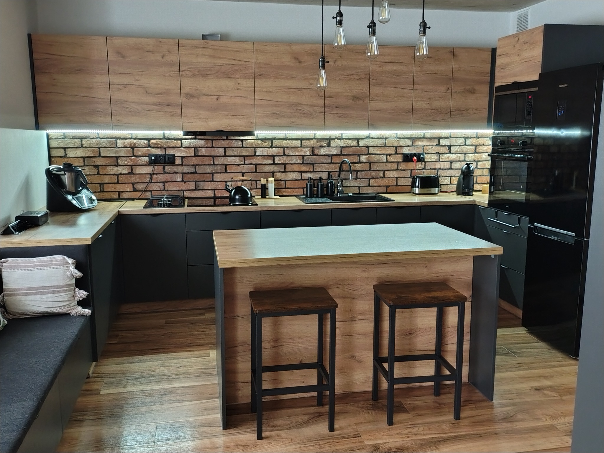 Nowoczesna kuchnia z wyspą: czarne matowe szafki dolne, drewnopodobne górne, ceglana ściana, oświetlenie LED, czarny sprzęt AGD i drewniana podłoga.