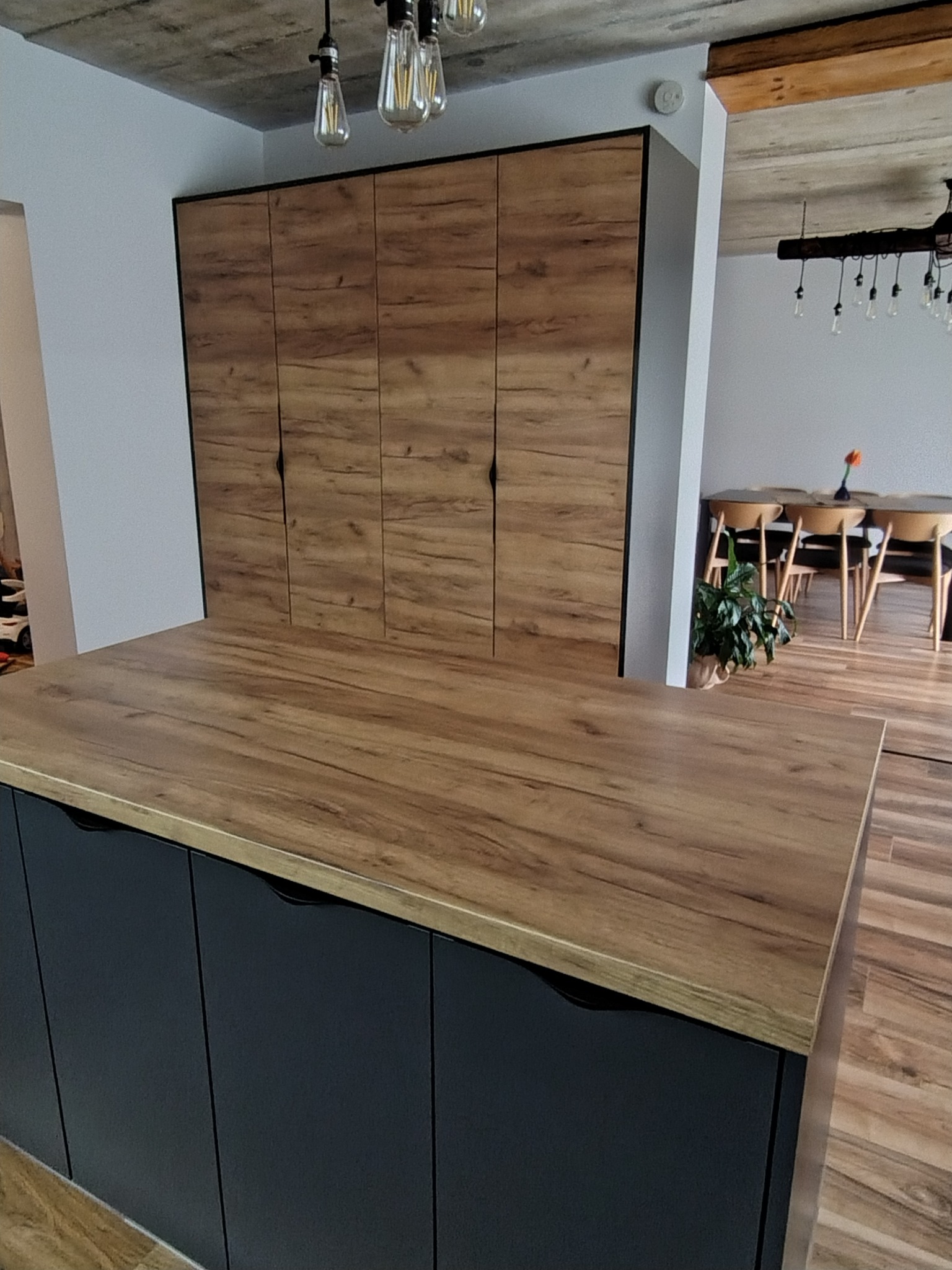 Nowoczesna szafa wnękowa z drewnianymi frontami i czarnymi uchwytami, widoczna z perspektywy blatu kuchennego, z elementami jadalni w tle.