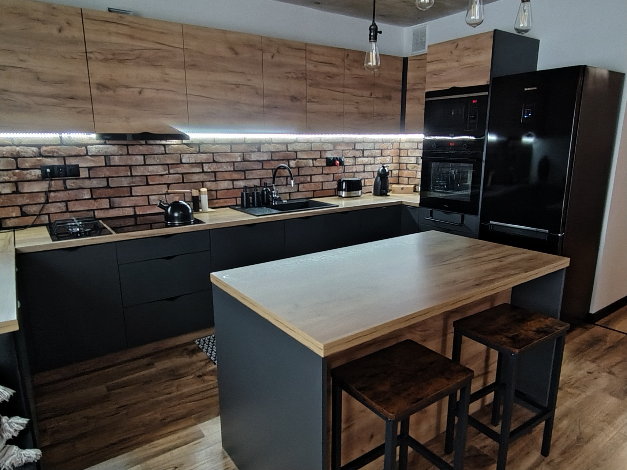Nowoczesna kuchnia z wyspą: czarne szafki, drewniane blaty i ceglana ściana. Uwagę przykuwają wiszące lampy w stylu industrialnym i drewniane hokery przy wyspie.