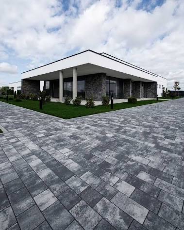 Nowoczesny dom z szarej kostki brukowej ułożonej w regularny wzór, trawnik i minimalistyczna architektura w tle. Kąt ujęcia podkreśla przestrzeń.