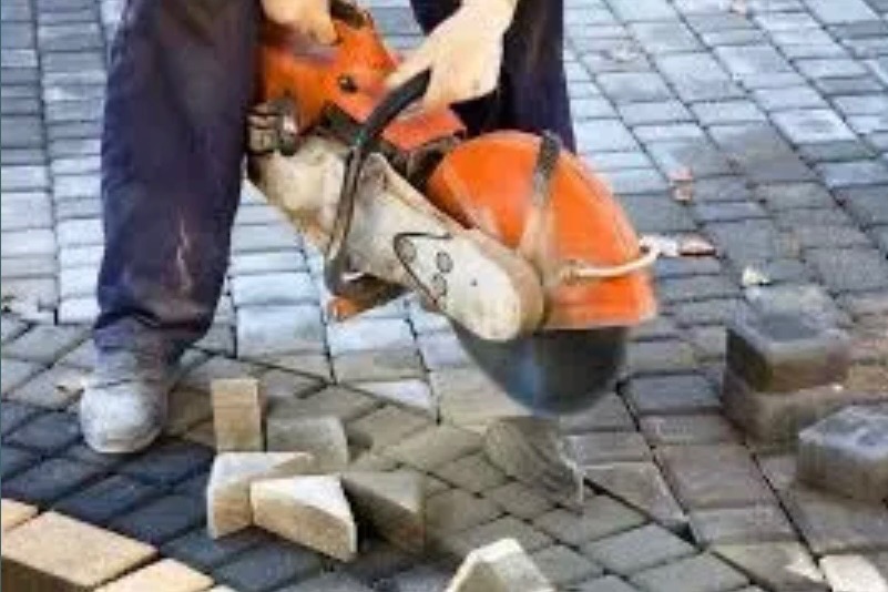 Pracownik przecina kostkę brukową szlifierką kątową, dopasowując ją do wzoru. Część kostki leży obok, tworząc stos. Widoczne ułożone fragmenty nawierzchni.