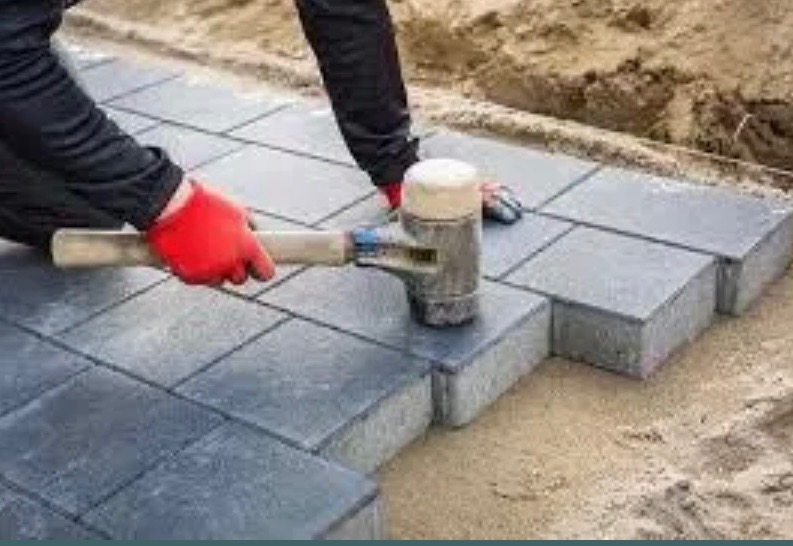 Układanie szarej kostki brukowej: pracownik w czerwonych rękawicach dobija gumowym młotkiem nową kostkę na piaskowym podłożu. Widoczny fragment ułożonej nawierzchni.