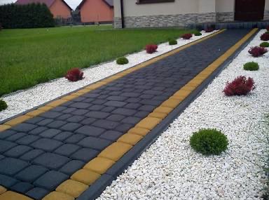 Długa, wybrukowana ścieżka z ciemnej kostki z żółtymi obrzeżami, otoczona białym żwirem i ozdobnymi krzewami prowadzi do domu z zielonym trawnikiem.