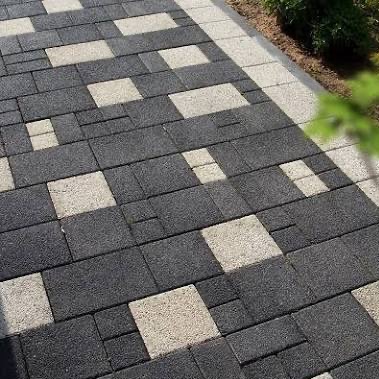 Wzór z czarno-białej kostki brukowej ułożonej na ścieżce, z bliska, z delikatnym zielonym akcentem rośliny w prawym górnym rogu i obrzeżem z jasnej kostki.