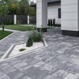 Szara kostka brukowa ułożona w geometryczny wzór z rabatą z białych kamieni i roślin, nowoczesny dom z kolumnami w tle. Oświetlenie ścieżki.