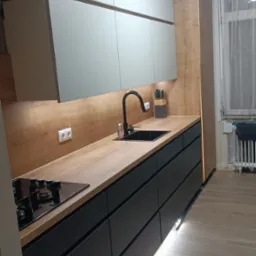 Nowoczesna kuchnia z szarymi szafkami górnymi i czarnymi dolnymi, drewniany blat i oświetlenie LED, widoczne poziomice pod szafkami.