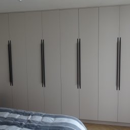 MBB-SYSTEM - Szafa wnękowa z jasnymi frontami i długimi, pionowymi uchwytami w sypialni. Widoczny fragment łóżka i okno.