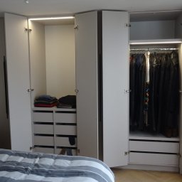 MBB-SYSTEM - Jasna szafa wnękowa z oświetleniem LED i szufladami, częściowo otwarta, ukazująca ubrania i złożone swetry, widok z sypialni.