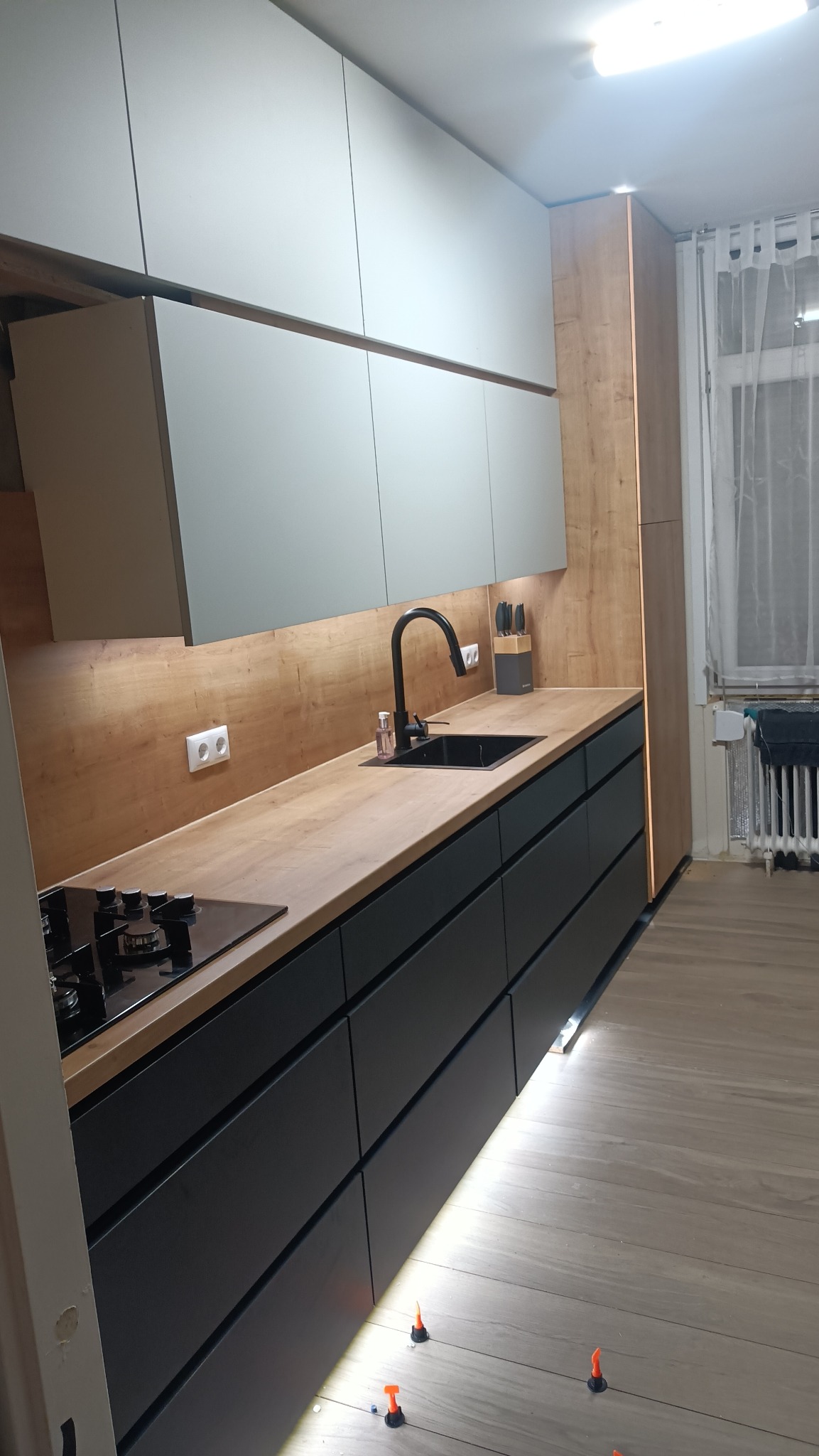 Nowoczesna kuchnia z szarymi szafkami górnymi i czarnymi dolnymi, drewniany blat i oświetlenie LED, widoczne poziomice pod szafkami.