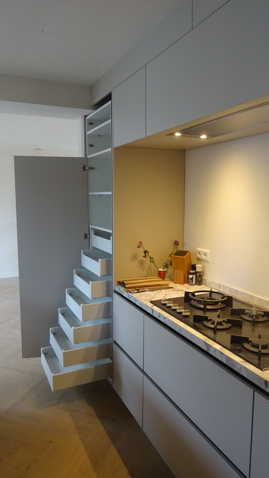 Nowoczesna kuchnia z szarymi szafkami i wysuwanymi szufladami w pionowej zabudowie. Blat marmurkowy, płyta gazowa.Minimalistyczny design z kwiatami.