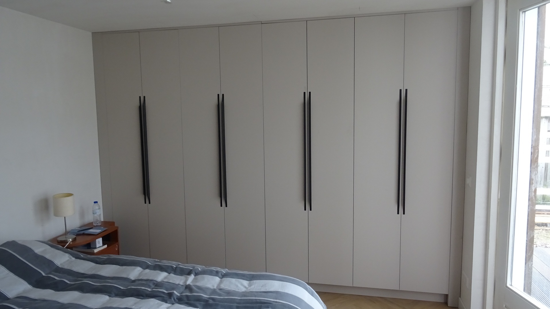Szafa wnękowa z jasnymi frontami i długimi, pionowymi uchwytami w sypialni. Widoczny fragment łóżka i okno.