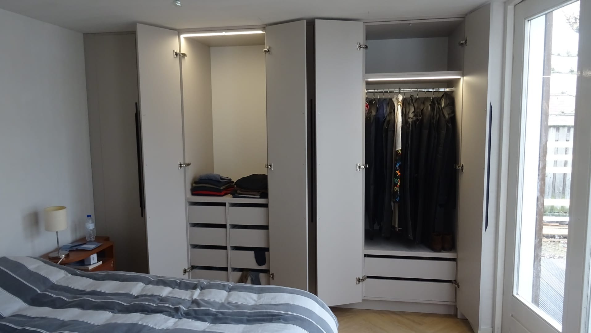 Jasna szafa wnękowa z oświetleniem LED i szufladami, częściowo otwarta, ukazująca ubrania i złożone swetry, widok z sypialni.