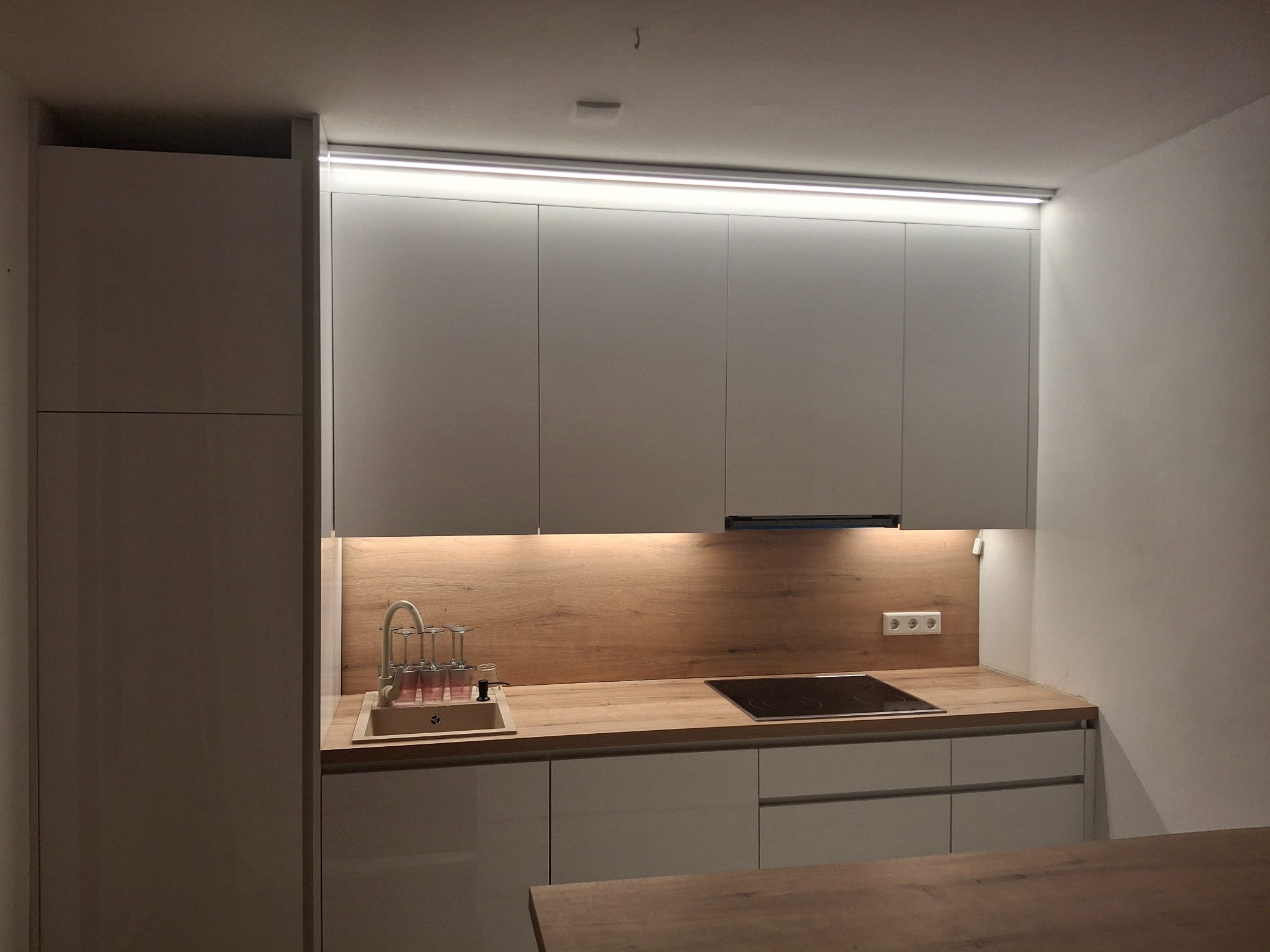 Nowoczesna kuchnia z szarymi frontami, drewnianym blatem i oświetleniem LED. Widoczny zlew z baterią, płyta indukcyjna i wiszące szafki.