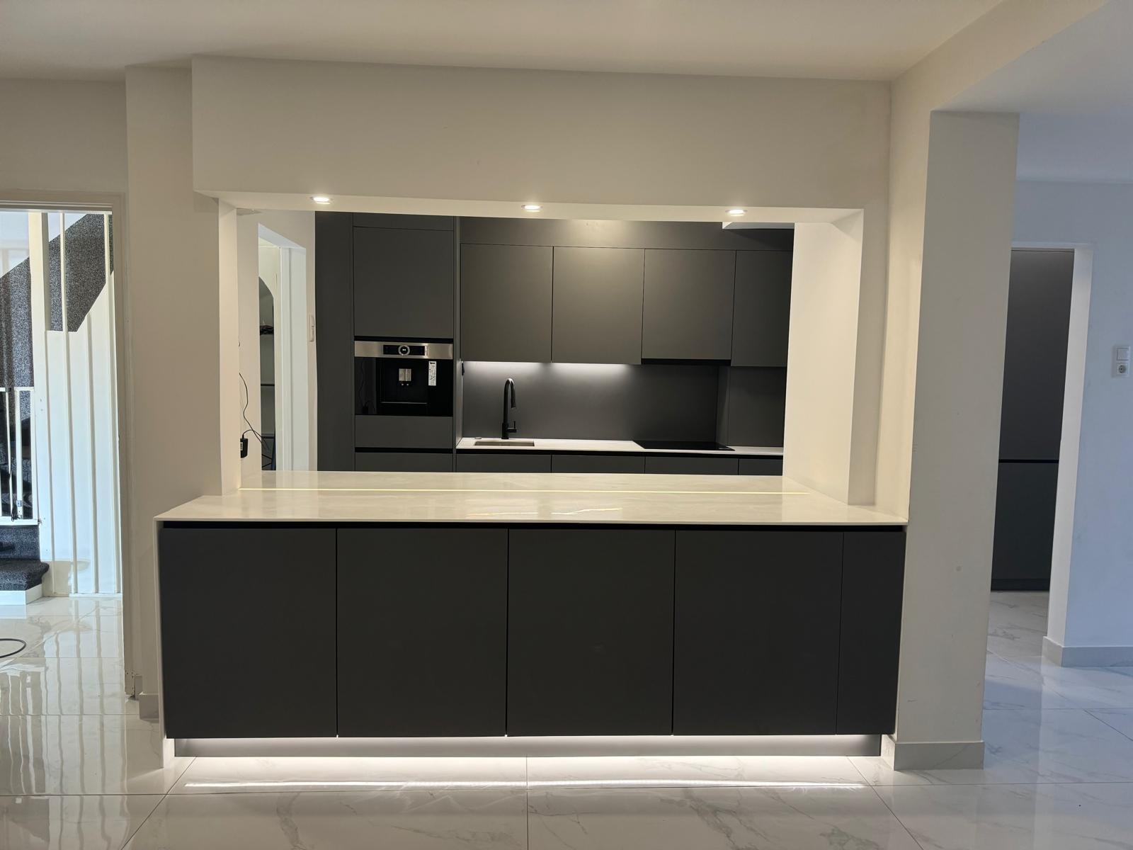 Nowoczesna kuchnia w odcieniach szarości z wyspą i subtelnym oświetleniem LED. Minimalistyczny design, zabudowa kuchenna z wbudowanym ekspresem do kawy. Marmurowe płytki podłogowe.