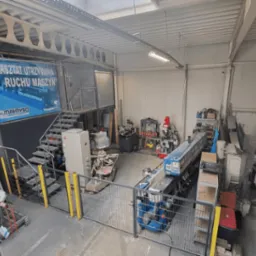 Widok z góry na przestronny warsztat z maszynami do obróbki metali, schodami i banerem reklamowym, oświetlony światłem dziennym.