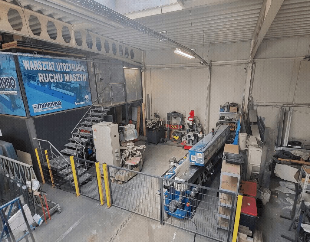 Widok z góry na przestronny warsztat z maszynami do obróbki metali, schodami i banerem reklamowym, oświetlony światłem dziennym.