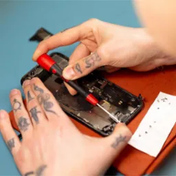 Rozłożony smartfon naprawiany precyzyjnym wkrętakiem Wiha w dłoniach serwisanta z tatuażami. Obok leżą małe śrubki.