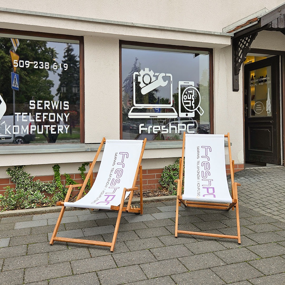 Fasada serwisu FreshPC z grafikami w oknach: laptop, telefon i narzędzia. Dwa leżaki z logo serwisu przed wejściem. Widoczny numer telefonu i godziny otwarcia.