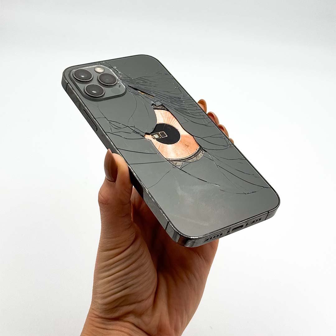 Dłoń trzyma uszkodzony, grafitowy iPhone 13 Pro Max z pękniętą obudową, ukazującą wewnętrzny moduł ładowania bezprzewodowego MagSafe. Widoczne pęknięcia na szkle.