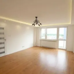 Jasne, przestronne wnętrze po remoncie: parkiet, białe ściany, okno z widokiem, designerska lampa i sufit z oświetleniem punktowym. Minimalistyczny styl.